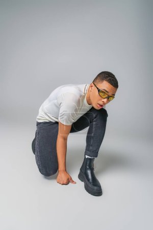 Foto de Hombre afroamericano de moda en camiseta, jeans y gafas de sol mirando hacia otro lado en gris - Imagen libre de derechos