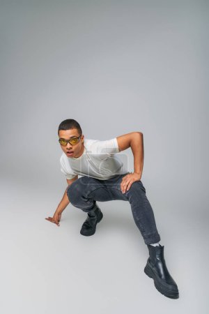 Photo for Modern style african american guy in t-shirt, jeans and sunglasses looking at camera on grey - Royalty Free Image