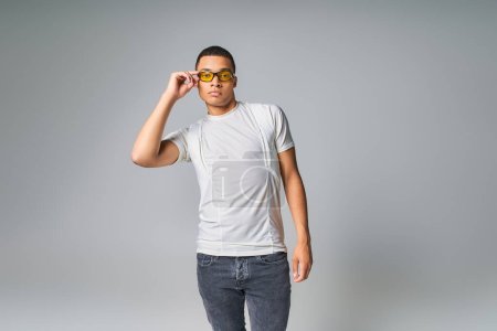 fashion trend, african american guy in t-shirt, jeans and sunglasses looking at camera on grey