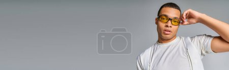 Photo for Trendy african american man in t-shirt adjusting sunglasses, looking at camera on grey, banner - Royalty Free Image