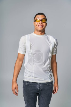 beeindruckte und gut gelaunte Afroamerikanerin in T-Shirt, Jeans, stylischer Sonnenbrille, die wegschaut auf grau