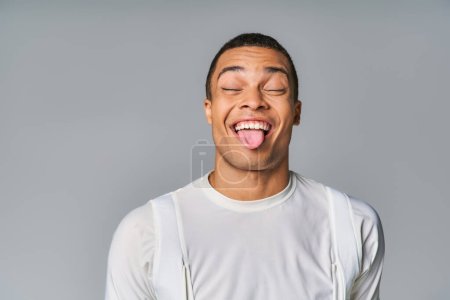 divertido hombre afroamericano en camiseta de moda, con los ojos cerrados, sobresaliendo sobre fondo gris