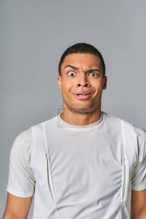 Téléchargez les photos : Surpris et découragé afro-américain en t-shirt élégant regardant la caméra sur gris - en image libre de droit