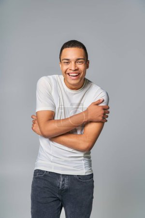 excited african american in stylish t-shirt looking at camera and laughing on grey