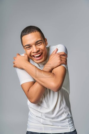 überglücklicher Afroamerikaner im trendigen T-Shirt blickt in die Kamera und lacht in grau