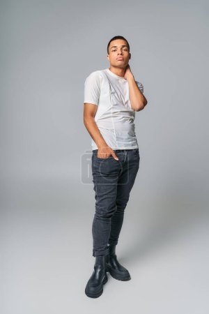 moderno afroamericano en camiseta y jeans, con la mano en el bolsillo, mirando a la cámara en gris