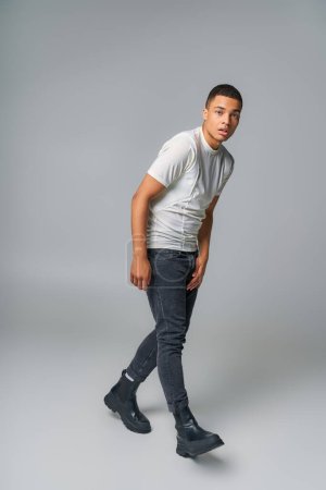 moderno afroamericano en camiseta y jeans, mirando a la cámara en gris, tendencia de la moda