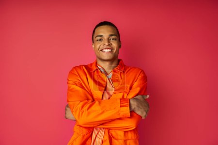 hombre afroamericano feliz en camisa naranja, con las manos dobladas, mirando a la cámara en rojo