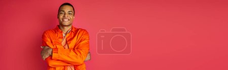 fröhlicher afrikanisch-amerikanischer Mann in orangefarbenem Hemd, mit gefalteten Händen, Blick in die Kamera auf rotem Banner