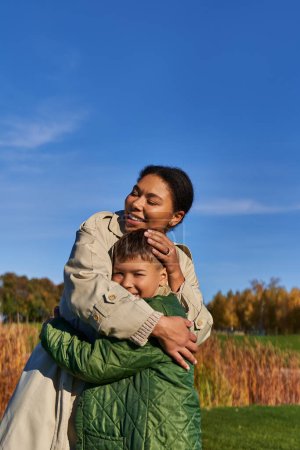 verbindendes, herbstliches Wesen, glückliche afrikanisch-amerikanische Mutter umarmt Sohn, Familie in Oberbekleidung, Herbst