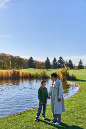 Photo for Family bonding, autumnal nature, african american mother walking with son along pond, hold hands - Royalty Free Image