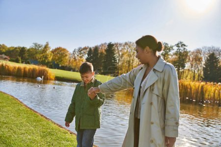 Photo for Family bonding, cheerful african american mother walking with son along pond, hold hands, autumn - Royalty Free Image