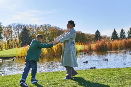 happy african american mother holding hands with son near pond with ducks, fall season, family