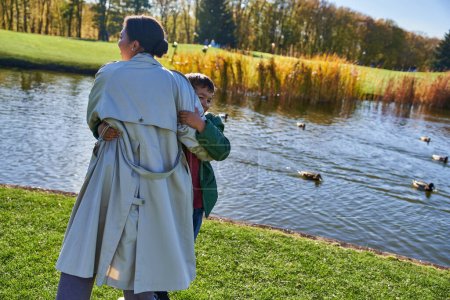 feliz afroamericano madre abrazando hijo cerca de estanque con patos, juguetón, temporada de otoño, alegría, familia