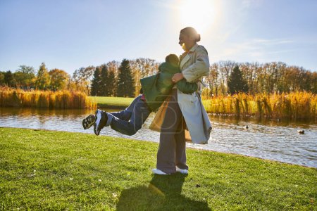 Téléchargez les photos : Franche, heureuse mère afro-américaine levant son fils ludique, s'amusant près de l'étang, vêtements de dessus, automne - en image libre de droit