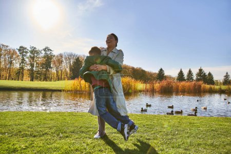 cándida, temporada de otoño, feliz madre afroamericana levantando a su hijo juguetón, divirtiéndose cerca del estanque