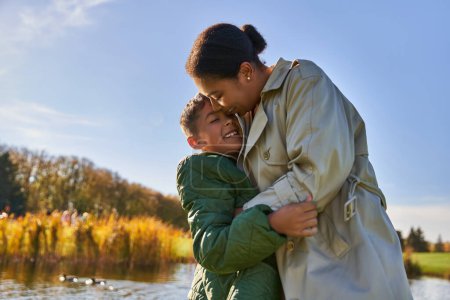 offen, Herbstzeit, fröhliche afrikanisch-amerikanische Mutter umarmt verspielten Sohn, hat Spaß in Teichnähe