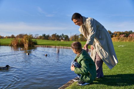 Herbst, fröhliche Afroamerikanerin in Oberbekleidung steht neben Sohn neben Teich mit Enten