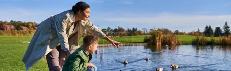 happy african american woman pointing at ducks in pond near son, autumnal nature, family, banner