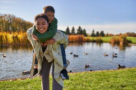positive mother piggybacking son near pond with ducks, childhood, african american, autumn, candid