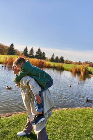 Herbstfarben, glückliche Mutter huckepack Sohn in der Nähe Teich mit Enten, Kindheit, afrikanisch-amerikanisch, Herbst