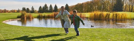 playful, happy african american mother and son running on grass near pond, modern parenting, banner