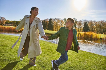 happy childhood, african american woman running with son near pond, outerwear, autumn, fall season