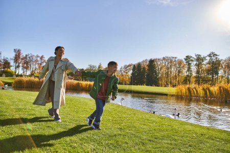 feliz infancia, mujer afroamericana en ropa de abrigo corriendo con hijo cerca de estanque, otoño, temporada de otoño