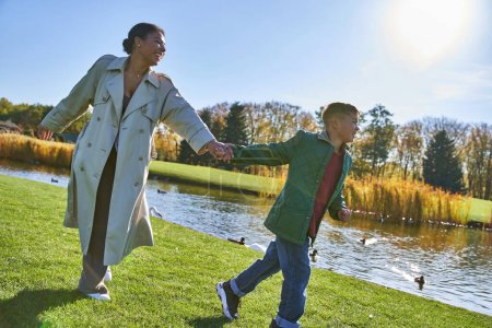 happy childhood, african american woman running with son near pond, autumnal outfits, fall season