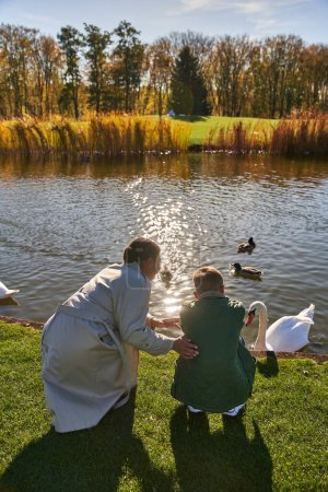 vista posterior de la mujer afroamericana y el niño mirando el lago con patos y cisnes, la infancia, la alegría