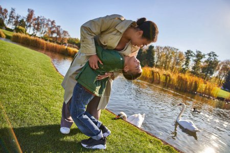 family love, happy african american mother and child having fun, hugging near lake, nature, autumn