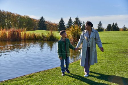 mother and son holding hands and walking on grass near lake, african american family, tranquil