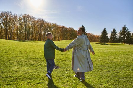 Journée ensoleillée, femme afro-américaine marchant avec son fils dans le parc, herbe verte, tenues d'automne, vêtements de dessus