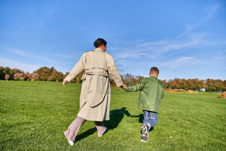 juguetona, mujer afroamericana corriendo junto con su hijo en el parque, día franco, soleado en otoño