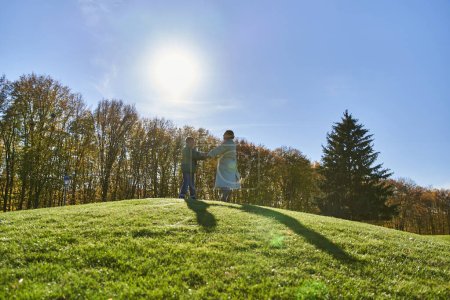 alegría, madre afroamericana y el niño tomados de la mano, de pie en la colina verde en el parque, follaje de otoño