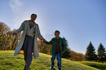 alegre africano americano madre y niño cogido de la mano, caminando por la colina, trajes de otoño