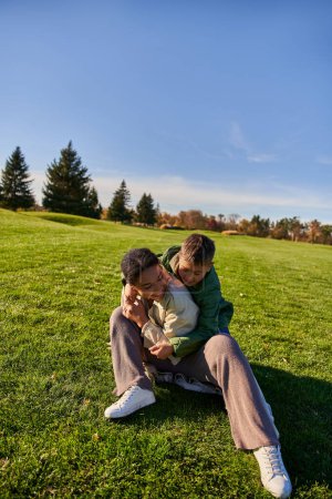 freudige Mutter und Sohn sitzen auf Gras, sonniger Tag, Herbst, verspielt afrikanisch-amerikanischer Junge umarmt Mutter