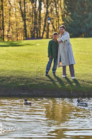 happy african american mother and son standing near lake, woman pointing at duck, nature, autumn