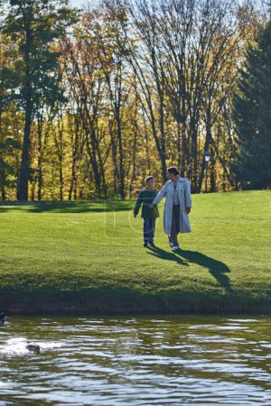 Photo for Happy african american mother and son walking towards lake, woman and boy in outerwear, nature, fall - Royalty Free Image