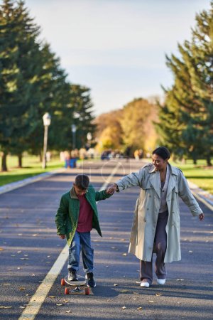 garçon chevauchant penny board et tenant la main avec sa mère, afro-américaine, feuilles d'or sur asphalte