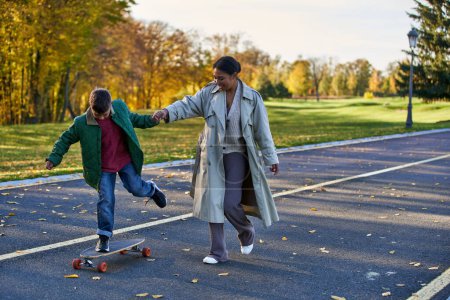 boy riding penny board and holding hands with happy mother, african american,  autumnal leaves
