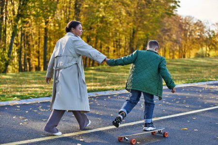 boy riding penny board and holding hands with cheerful mother, african american,  autumnal leaves