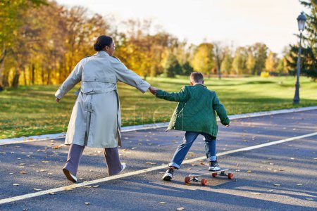 Junge in Oberbekleidung reitet Penny Board und hält Hand an Mutter, afrikanisch-amerikanisch, Herbstblätter