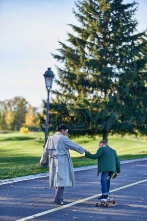 Photo for Kid in outerwear riding penny board and holding hands with mom, african american,  autumn leaves - Royalty Free Image