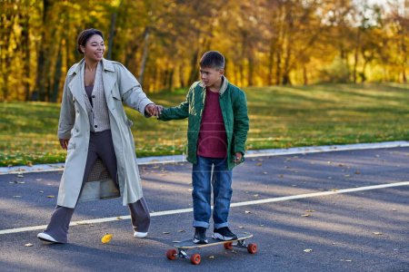 Kind in Oberbekleidung reitet Penny Board und hält Händchen mit glücklicher afrikanisch-amerikanischer Mutter, Herbst