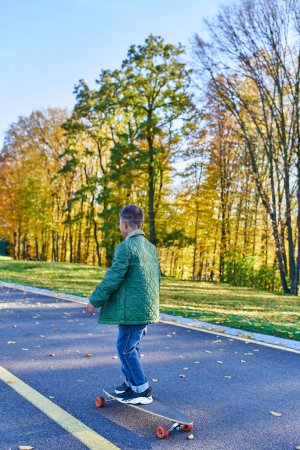 niño en ropa de abrigo otoñal a caballo penny board, asfalto, parque en temporada de otoño, hojas de oro, lindo niño