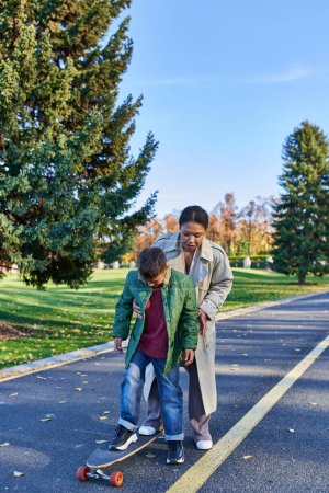 mother and son in autumn park, african american woman teaching boy how to ride penny board, active