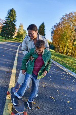 Mutter und Sohn im Herbst Park, afrikanisch-amerikanische Frau unterstützt Jungen auf Penny Board, freudige Momente
