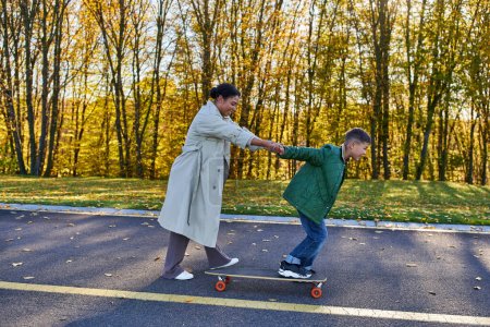 Téléchargez les photos : Mère et fils dans le parc d'automne, heureuse femme afro-américaine tenant la main avec le garçon sur la planche de penny - en image libre de droit