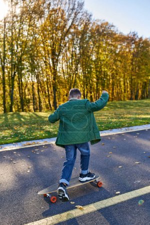 back view of boy in outerwear and jeans riding penny board in park, autumn, golden leaves, cute kid